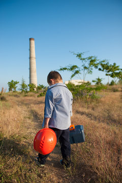 sad little caucasian boy with helmet and tool case in hands walking towards factory with pipe