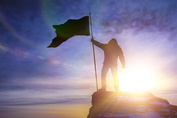 silhouette of man with flag of victory on top of a mountain over sky and sun light background....