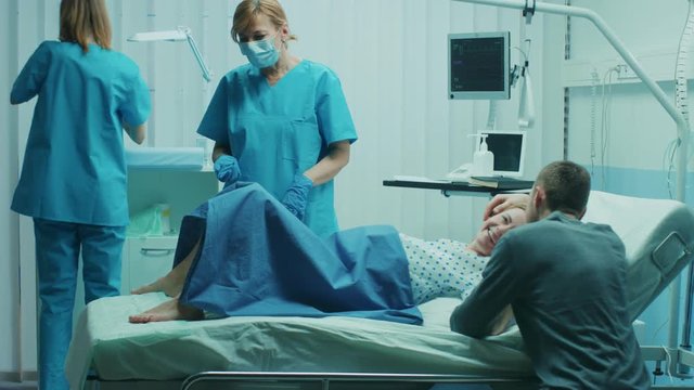In the Hospital Woman in Labor Pushes to Give Birth, Baby Comes out, Obstetricians Assist Delivery, Husband Holds Supports His Wife. Side View Footage. Shot on RED EPIC-W 8K Helium Cinema Camera.