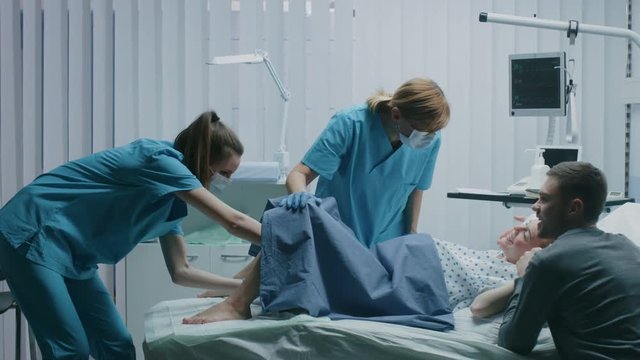 In the Hospital Woman in Labor Pushes to Give Birth, Baby Comes out, Obstetricians Assist Delivery, Husband Holds Supports His Wife. Side View Footage. Shot on RED EPIC-W 8K Helium Cinema Camera.