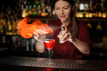 Young female bartender sprinkling a cocktail glass filled with Aperol syringe cocktail with a peated whisky and making a smoky note