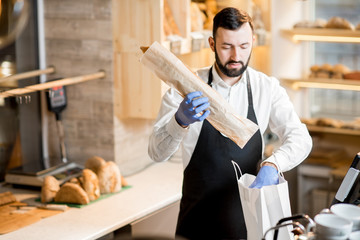 Bread seller packing a baguette into the paper bag in the store with bakery products