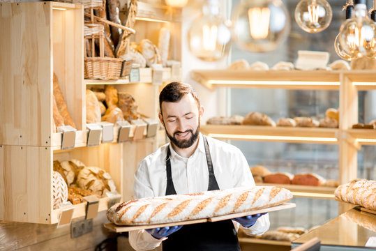 Handsome seller in uniform holding a big loaf of bread in the store with bakery products