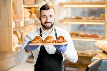 Portrait of a handsome seller in uniform standing with delicious croissants in the store with...