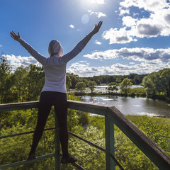 молодая спортивная женщина в спортивной одежде и толстовке с капюшоном стоит спиной к камере, лицом к солнцу с распростертыми руками на высоте в парке 