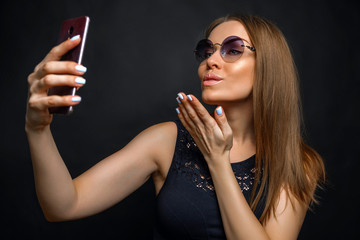 красивая молодая женщина держит смартфон в руке и делает селфи с воздушным поцелуем в солнечных очках и черном платье на черном фоне 