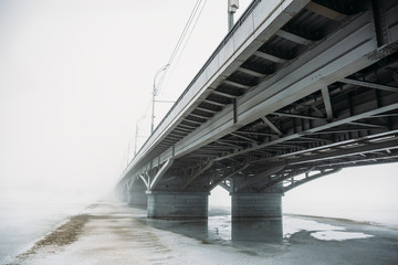 Bridge in fog over frozen river in ice and snow. Mysterious urban landscape with mist