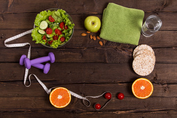 Obraz na płótnie Canvas proper nutrition, fitness food, water, fruit, green apple, kiwi, orange, vegetable salad. program of diet, dumbbells. The concept of healthy food on a dark wooden background for advertising your text