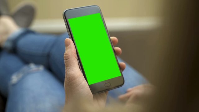 Female holds and reads her smartphone in living room with green screen / chroma keyed smartphone. 