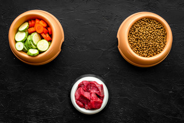pet care with dry food, vegetables and raw meat for dog or cat in plastic bowl on black background top view space for text