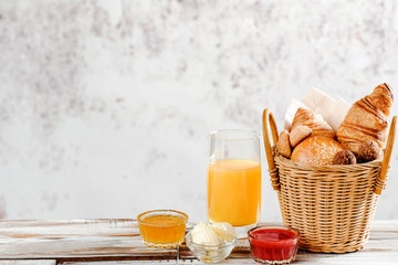Baking breakfast croissants and bun in basket with orange juice, honey, butter, raspberry jam on white wood table