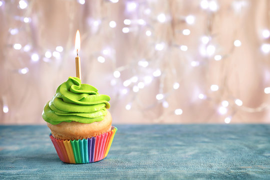 Birthday cupcake with burning candle on table against blurred lights