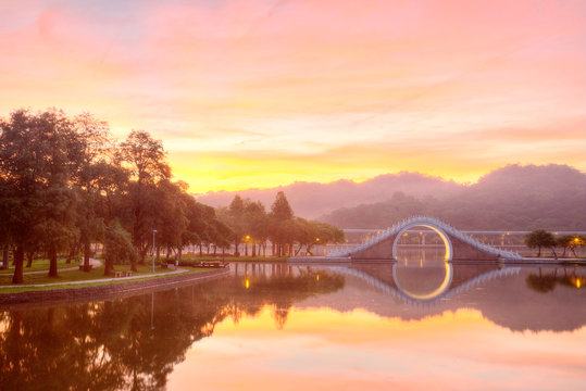 Scenery of an urban park lit up by golden sunlight with reflections of an arch bridge and glowing sky on the smooth lake water ~ Early morning scene of Dahu Community Park in Nahu,Taipei City Taiwan