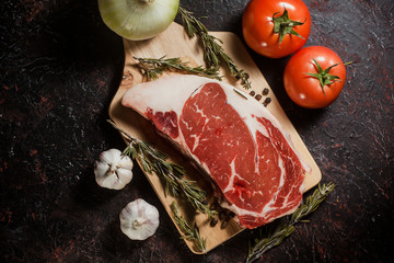 Raw ribeye steak garnished with a sprig of rosemary, garlic and onion and tomatoes