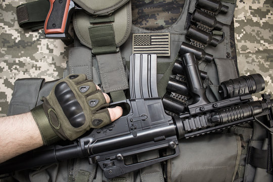 Upper view photo of hand in tactical gloves holding a rifle on military tactacal bulletproof vest, cartrige belt laying on camouflage cloth background.