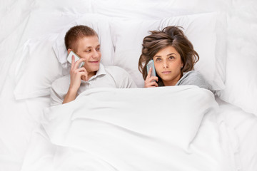 Concentrated young woman and funny man lying in bed and talking on phones