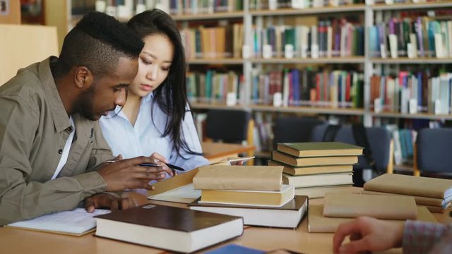 Young african american and asian college students working together preparing for exams while sitting at table at university library with classmates