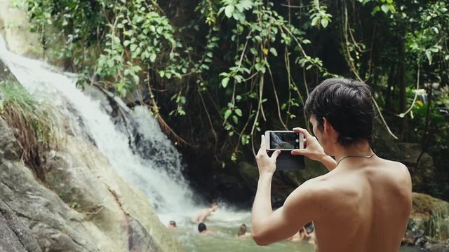 Young attractive man taking photo with beautiful waterfall and swimming people in water on the background. slow motion. 1920x1080