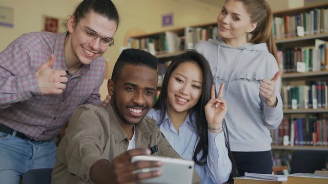 Group of international students have fun smiling and making selfie photos on smartphone camera at university library. Cheerful friends have rest while preapre project together