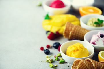 Ice cream balls in bowls, waffle cones, berries, orange, mango, pistachio on grey concrete background. Colorful collection, flat lay, summer concept