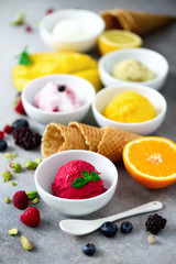 Ice cream balls in bowls, waffle cones, berries, orange, mango, pistachio on grey concrete background. Colorful collection, flat lay, summer concept