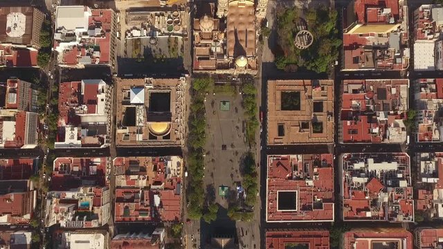 Overhead aerial view straight down onto the plazas and red tiled blocks of the old centre of Guadalajara in Mexico with domes and courtyards below. A vivid and elegant scene of the city grid.
