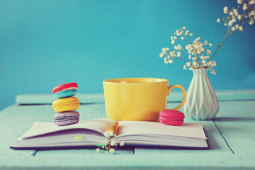 Big yellow cup of tea with flower and macaroons and notebook on blue background. Toned picture