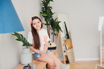 happy preteen girl resting at home, sitting with cup of hot tea with houseplants on backgroung in...