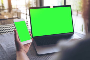 Mockup image of a woman holding blank mobile phone while using laptop with blank green screen on table - Powered by Adobe