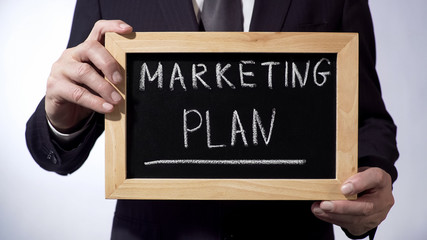Marketing plan written on blackboard, man in black suit holding sign, business