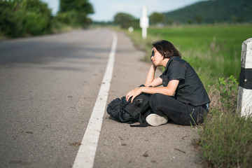 Woman sit with backpack hitchhiking along a road in countryside