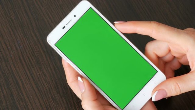 Female hands with beautiful French manicure take a white smartphone with Green Screen. Using Smartphone,Holding Smartphone with Green Screen on a stylish black wooden table