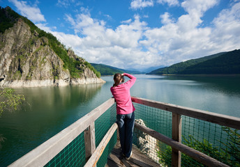 Rear view of female photographer on the lake Vidraru Carpathians Romania. Beautiful scenery of a sunny day: mountains, forests, lake, blue sky with clouds
