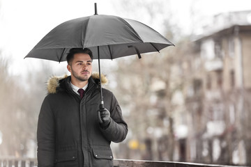Young man in warm clothes with dark umbrella outdoors
