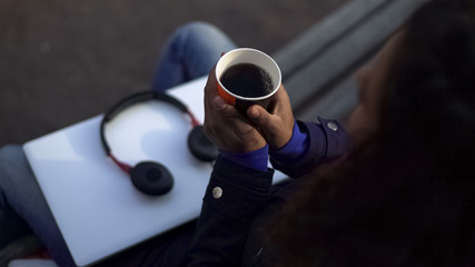 Woman warming hands, holding cup with fragrant hot drink while sitting outdoors