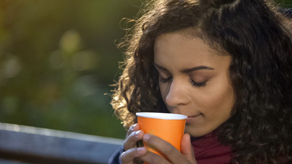 Young curly-haired woman enjoying taste of favorite morning coffee outdoors