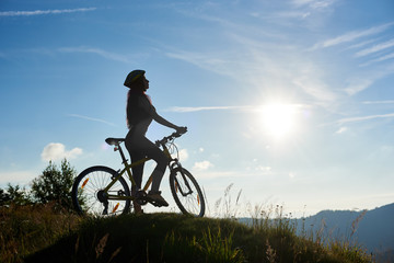 Fototapeta na wymiar Silhouette of sporty girl cyclist riding on bicycle in the mountains, wearing helmet, enjoying sunrise on sunny morning. Outdoor sport activity, lifestyle concept. Copy space