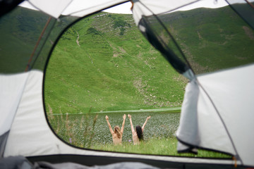 Back view of two attractive naked women lifting hands up in the air near the lake in the mountains....