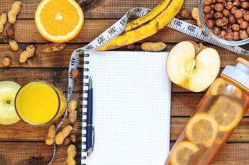 Healthy lifestyle; fitness bottle; fruits (oranges; apples and bananas); hazelnuts and peanuts; orange juice, a pen and a notebook and measuring tape. Top view; the concept of free space for text