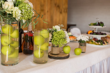 Rustic flower arrangement with white flowers and greenery in a glass vase with water and apples at a wedding banquet. Table set for an event party or wedding reception.