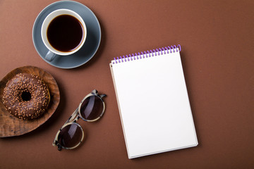 Obraz na płótnie Canvas Chocolate donuts with cup of black coffee, empty notebook with text space and sun glasses on brown background. Monochromatic concept.