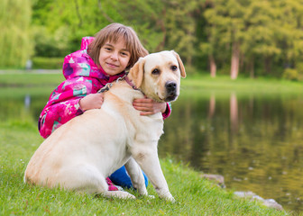 Little girl with labrador retriever on walk in park. Child sitting on green grass with dog - outdoor in nature portrait. Pet, domestic animal and people concept.