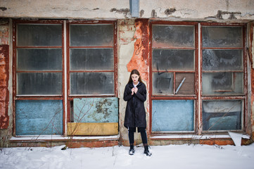 Fashionable long legs brunette model in long black cloak posed outdoor at winter day against old grunge wall with broken windows.