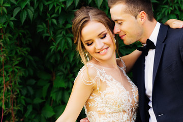 smiling bride with a delicate makeup hugs her lover who closed his eyes near green bushes