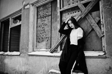 Fashionable long legs brunette model in long black cloak posed outdoor at winter day against old grunge wall with broken windows.