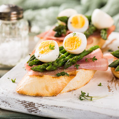 Easter breakfast or lunch with sendwich with ham, asparagus and quail eggs on white old chopping board on wooden background. Spring food concept.