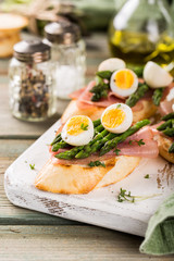 Fresh sendwich with ham, asparagus and quail eggs on white old chopping board on wooden background. Easter spring breakfast concept with copy space.