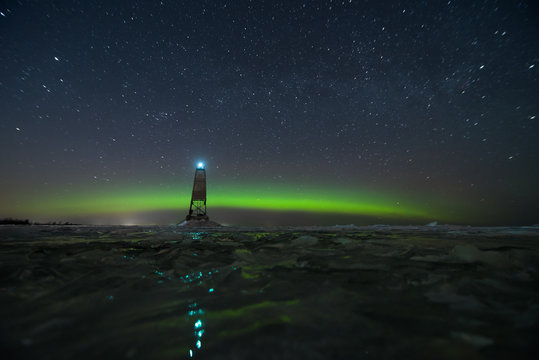 Wide panoramic night sky view of milky way and beautiful green aurora borealis (northern polar lights) in Finland with reflection on the ice floes on frozen lake with lighthouse on the shore