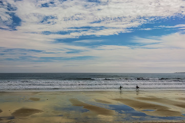 the shore of the ocean at low tide, the reflection of clouds in the surface of sand and water, two serfengists with boards go in waves along the shore.  Atlantic Ocean. USA
