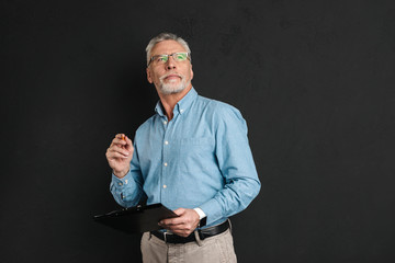 Portrait of middle aged man 60s with grey hair and beard looking upward while holding clipboard with documents, isolated over black background